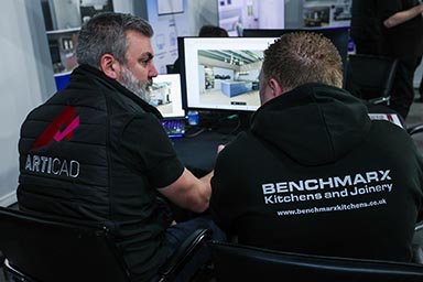 Two men, one with an ArtiCAD jacket and another with a Benchmarx jacket, sitting in front of a computer.