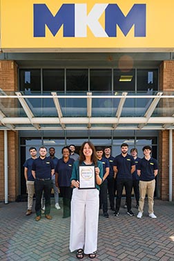 A woman holding a certificate in front of a group of people in matching T-shirts at the front of an MKM building.